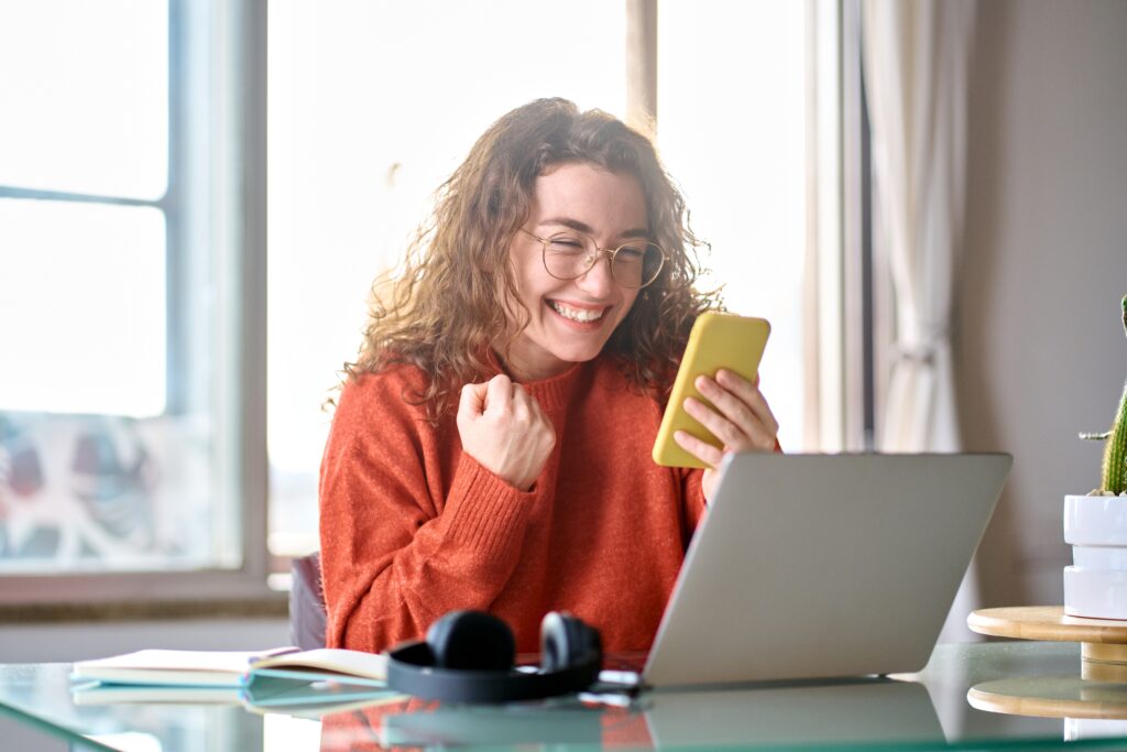 Woman wearing glasses and a red sweater sitting in front of a laptop and holding her phone, pumping her fist victoriously.