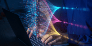 Gros plan d'une paire de mains en train de taper sur un ordinateur portable. Un hologramme de plusieurs lignes bleues, roses et jaunes relie des chaînes de code binaire à un ensemble de binaires de ces couleurs qui flottent au-dessus du clavier.